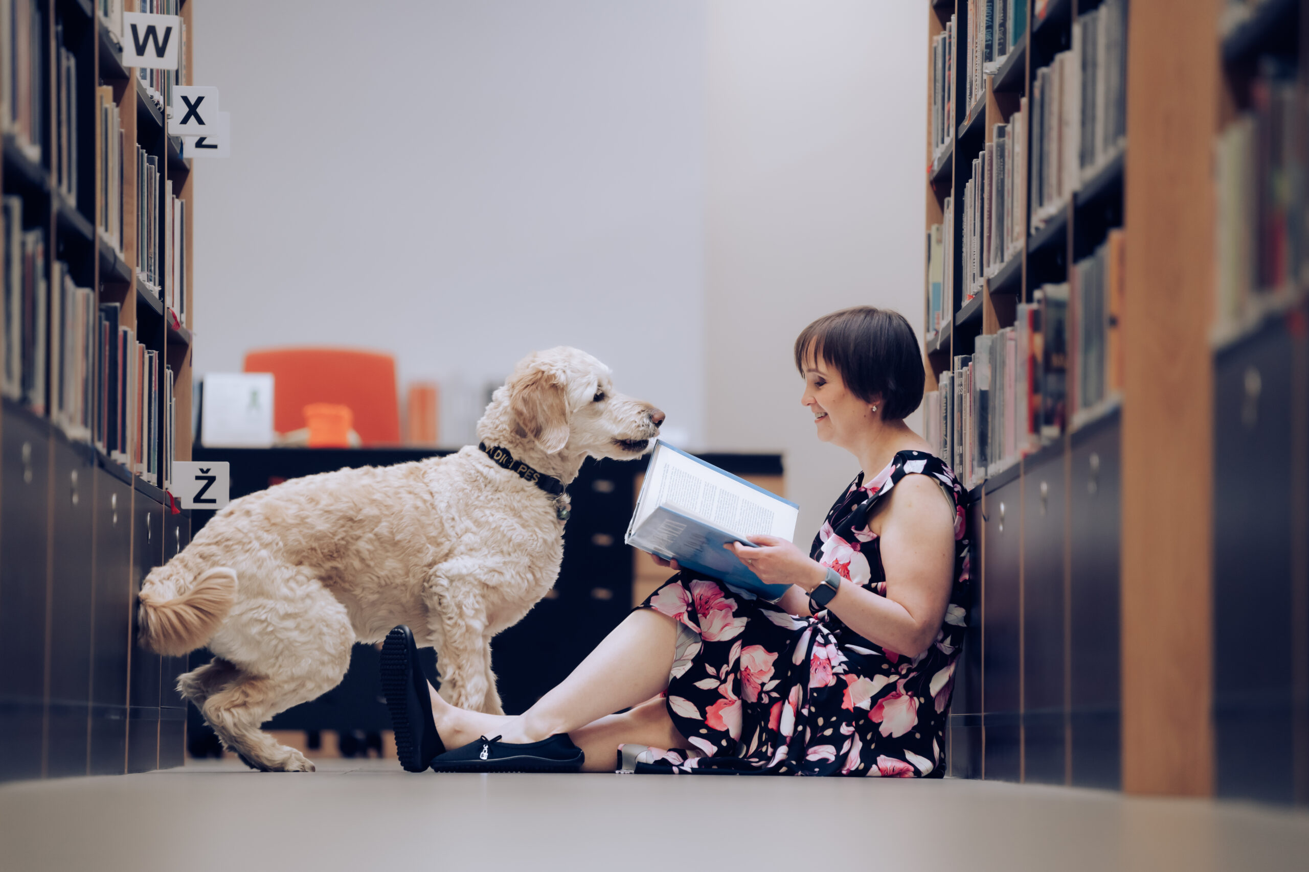 Spisovatelka Anna Burdová sedí v knihovně na zemi, opírá se o regál s knihami, drží knihu a před ní je vodicí fenka Dixie s dychtivým výrazem.